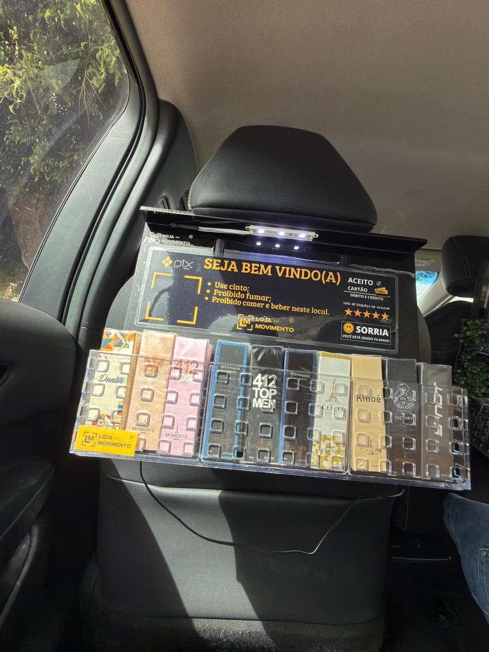 Lojinha de perfumes instalada no encosto do banco da frente de um carro, com bandeja de acrílico transparente cheia de perfumes de bolso e placa de boas-vindas ao passageiro.