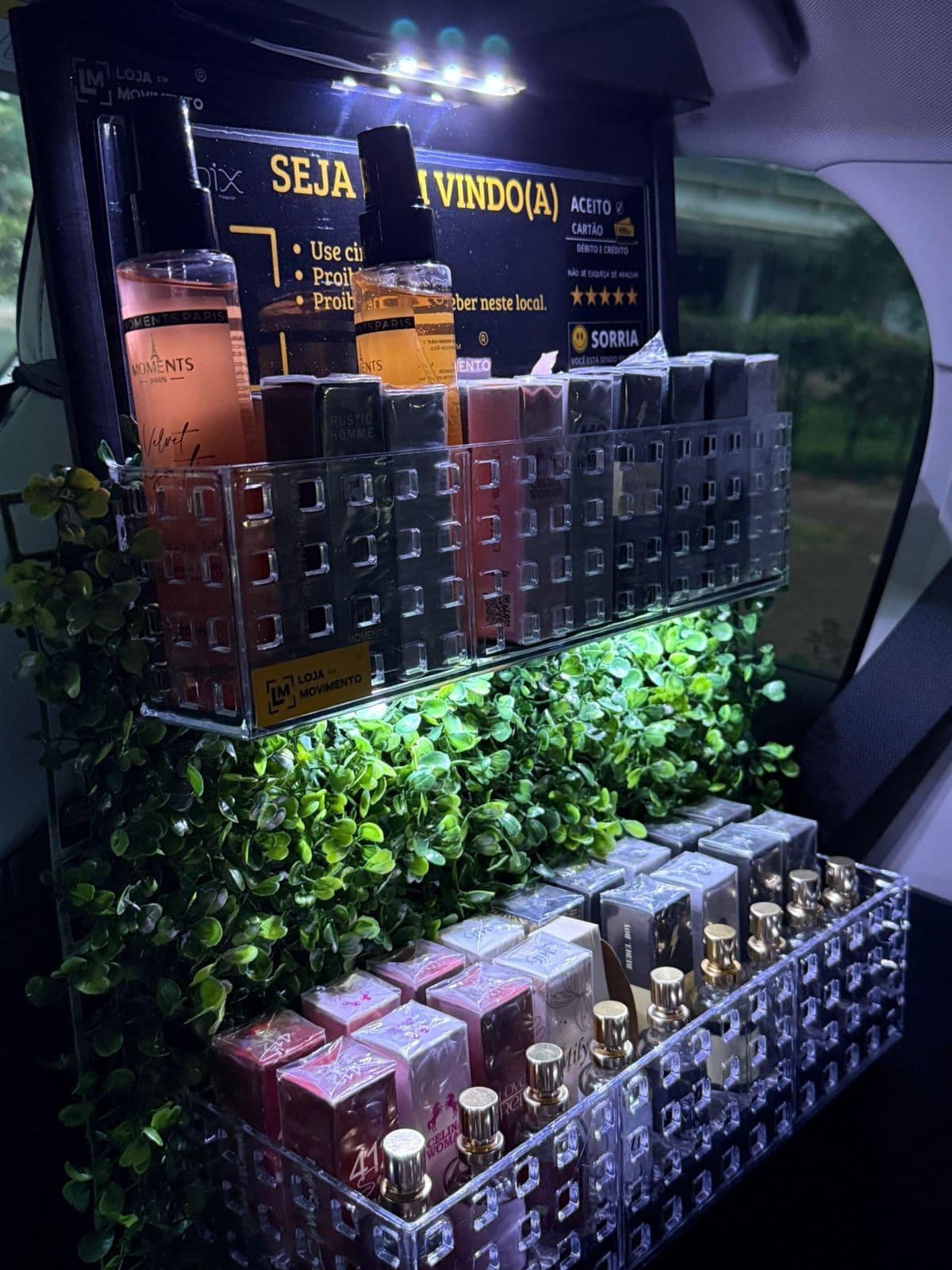 Lojinha de perfumes montada no banco de trás de um carro à noite, com bandejas de acrílico iluminadas por LED e dezenas de perfumes de bolso Moments Paris expostos.